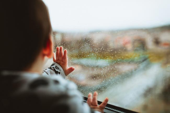 Niño pequeño retratado de espaldad con una mano apoyada en una ventana